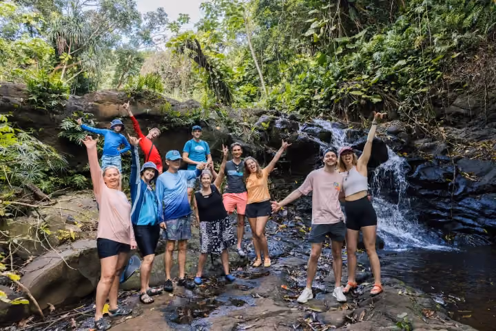 Travelers pose beside a cascading waterfall in a lush tropical jungle.