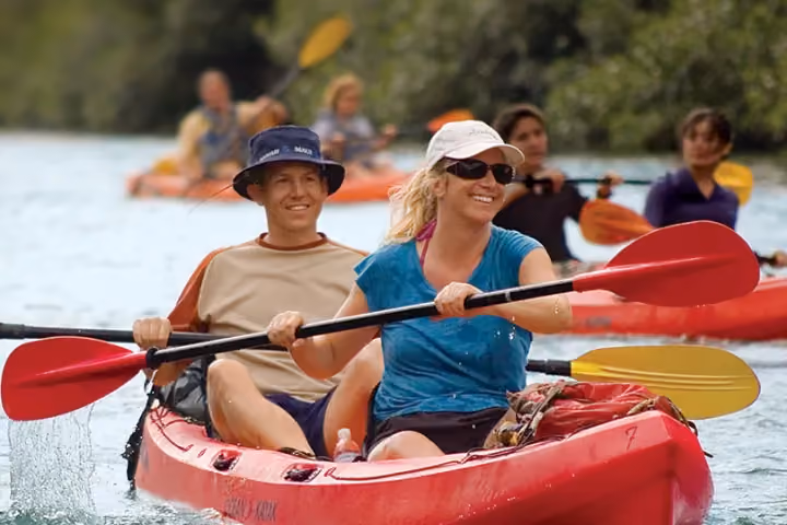 Smiling couple paddles a red tandem kayak on calm water.