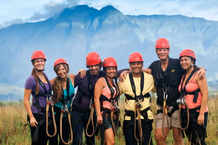 Seven smiling guests in red helmets and harnesses pose before green mountains.