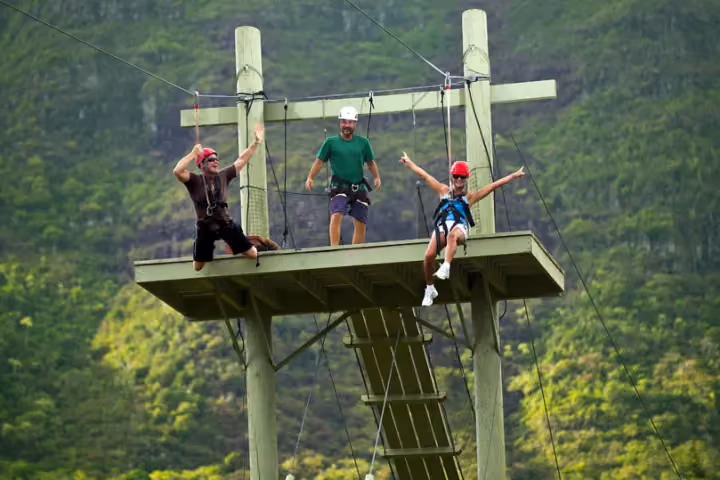 Guests and a guide sit harnessed on a wooden zipline platform against forested hills.