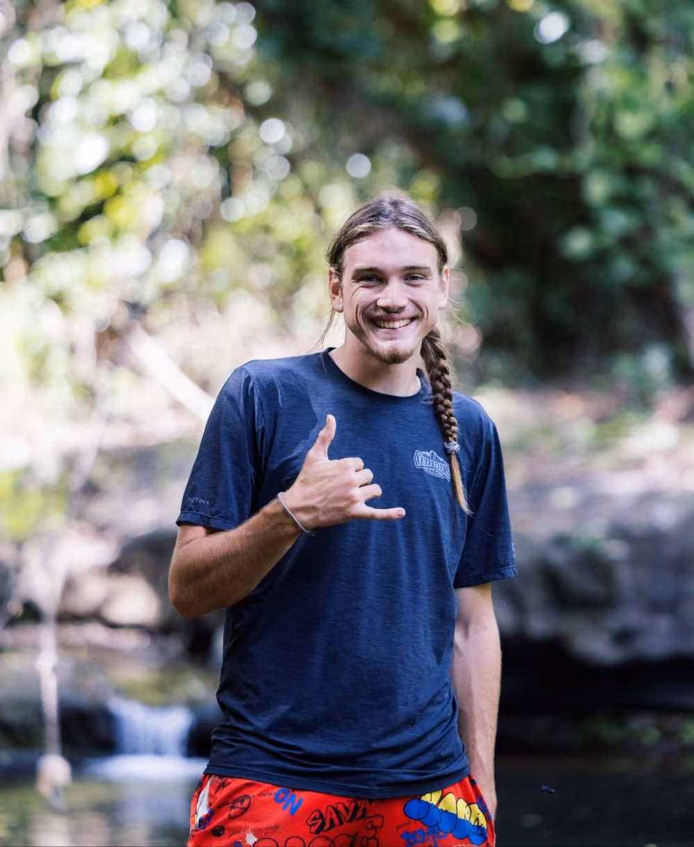 Outfitters Kauai guide smiling and throwing a shaka near a waterfall on the Zipline Safari and Huleia River adventure
