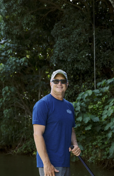 Man in a blue shirt and cap holding a paddle beside green foliage