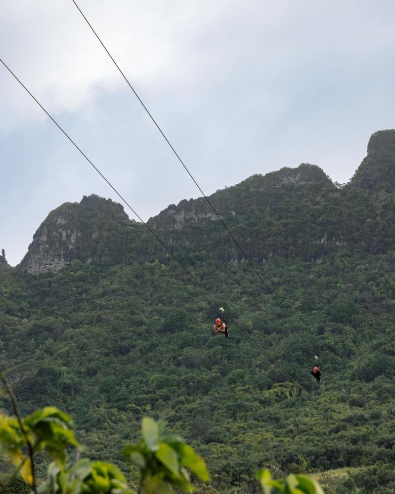 Guests ziplining across lush green valleys at Kīpū Ranch on Kauaʻi, with the Hāʻupu Mountain Range in the background