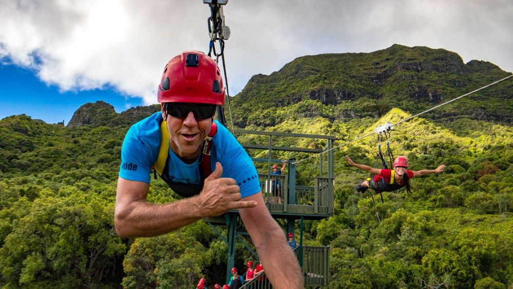 Smiling guest giving a shaka on a zipline with another rider and the platform behind
