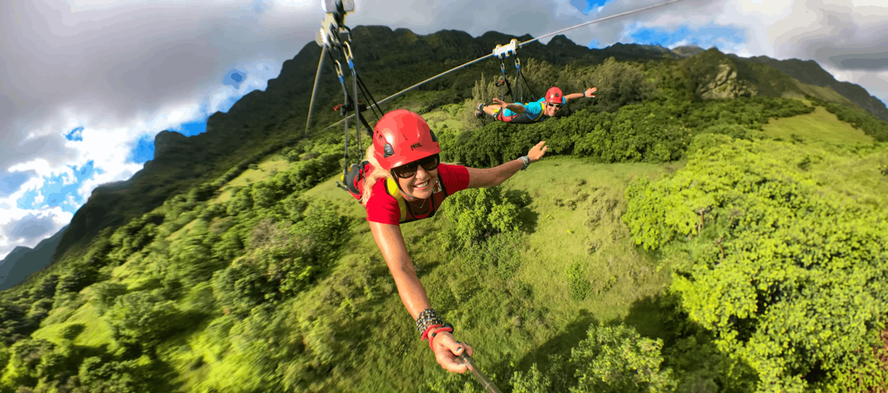Zipliner in a red helmet flying headfirst on the FlyLine over Kīpū Ranch with a second rider behind