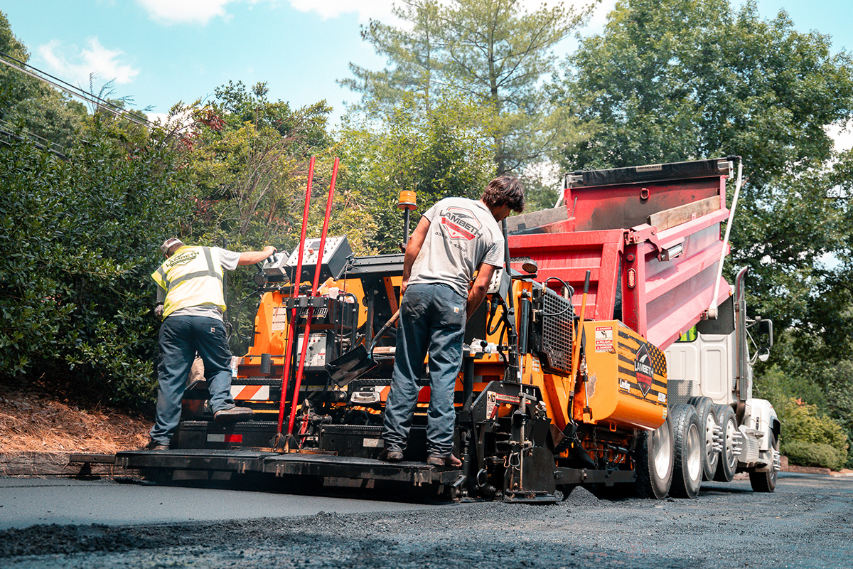 Lambeth Asphalt Services crew members operating a paver and dump truck along a wooded driveway during paving work at Friendly West in Greensboro, NC