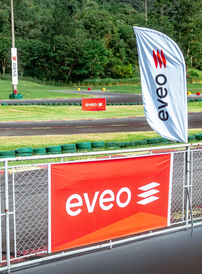 Bandeiras e banner da EVEO ao redor de uma pista de kart, com barreira de pneus e mata ao fundo