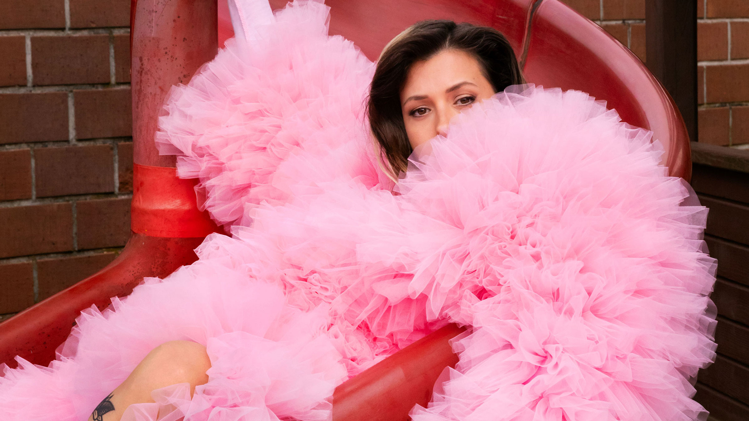 Frau mit braunen Haaren in voluminösem rosa Tüllkleid sitzt in roter Rutsche neben Backsteinwand.