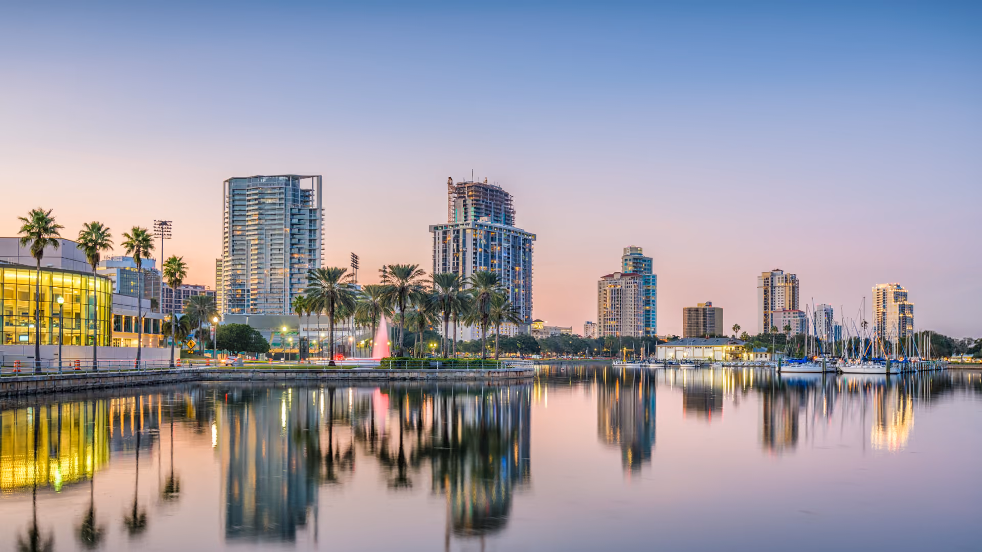 Calm St Petersburg waterfront with palm trees, high-rise buildings, cranes, and boats reflected at sunset.