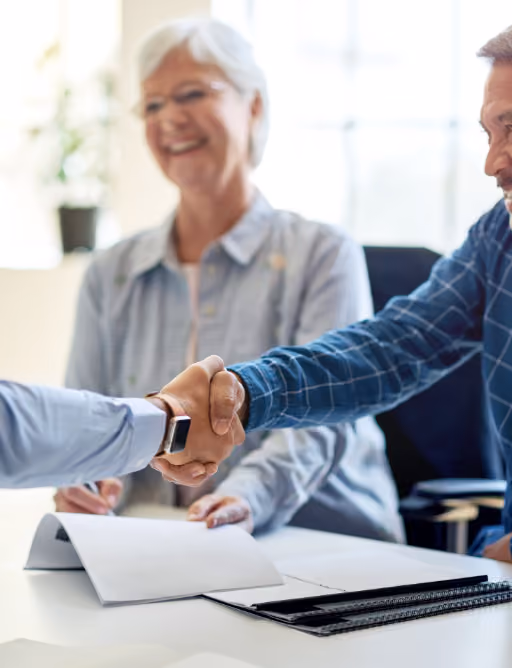 Two men shaking hands over a table with documents, a woman smiling in the background.