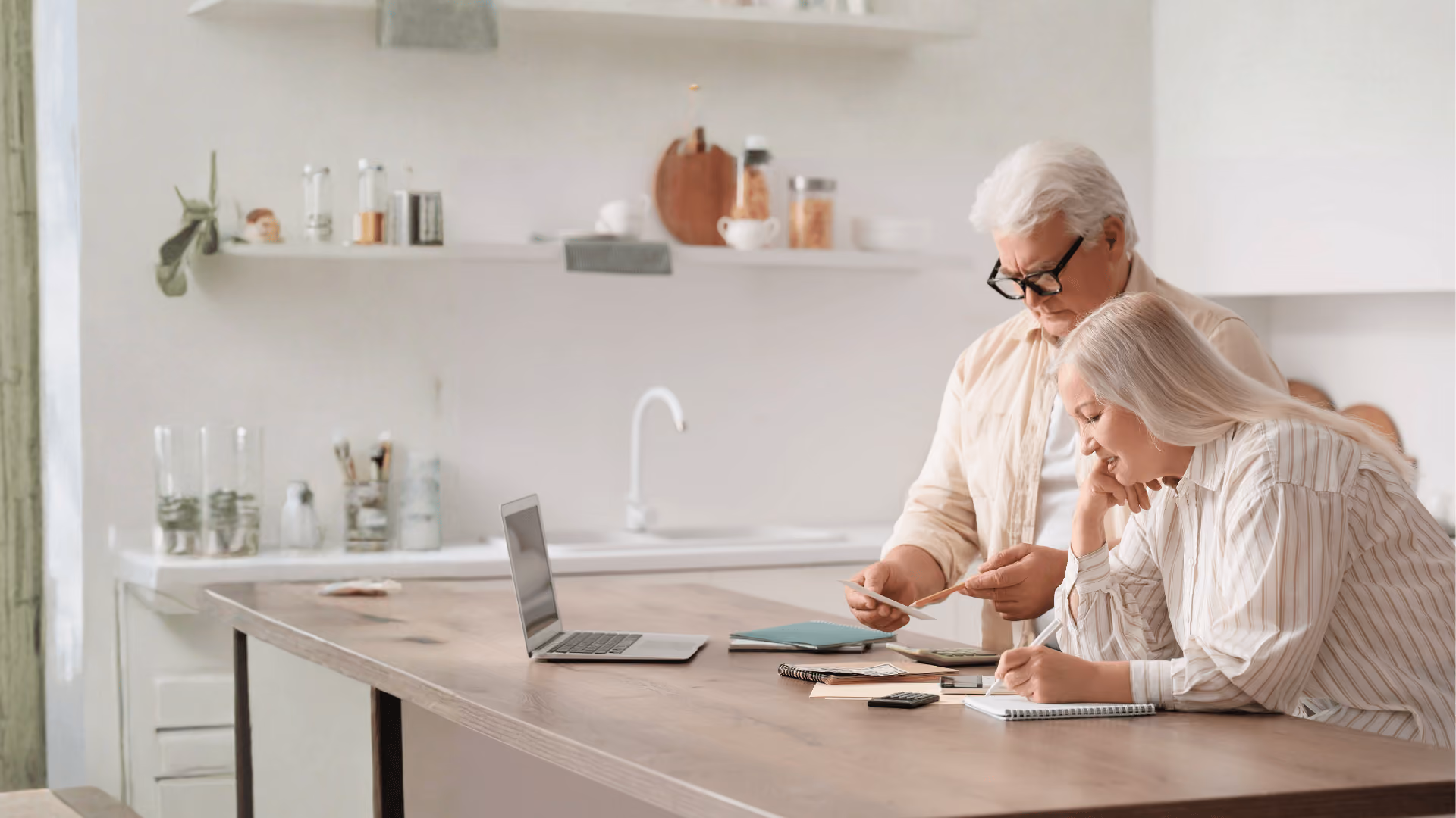 Senior couple working together at kitchen table with laptop, documents, and calculator.