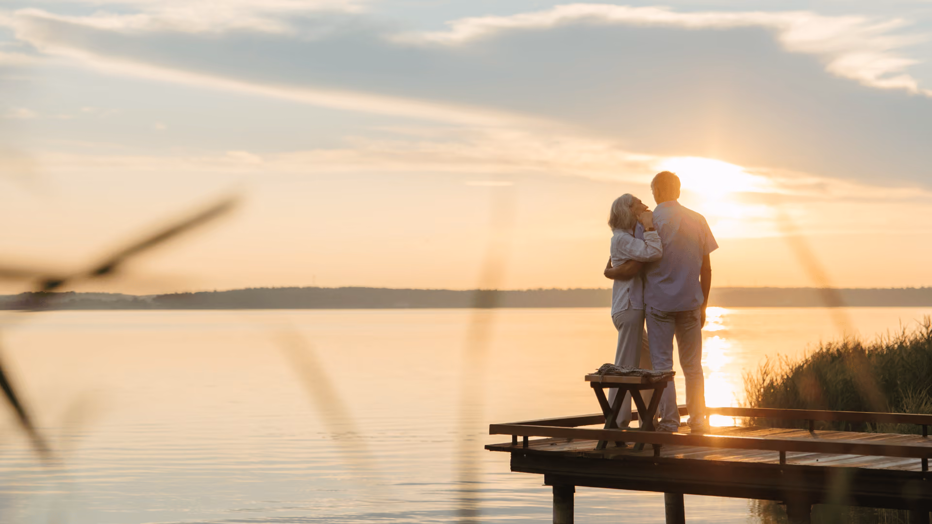 Older couple embracing on a dock at sunset by a calm lake with soft clouds