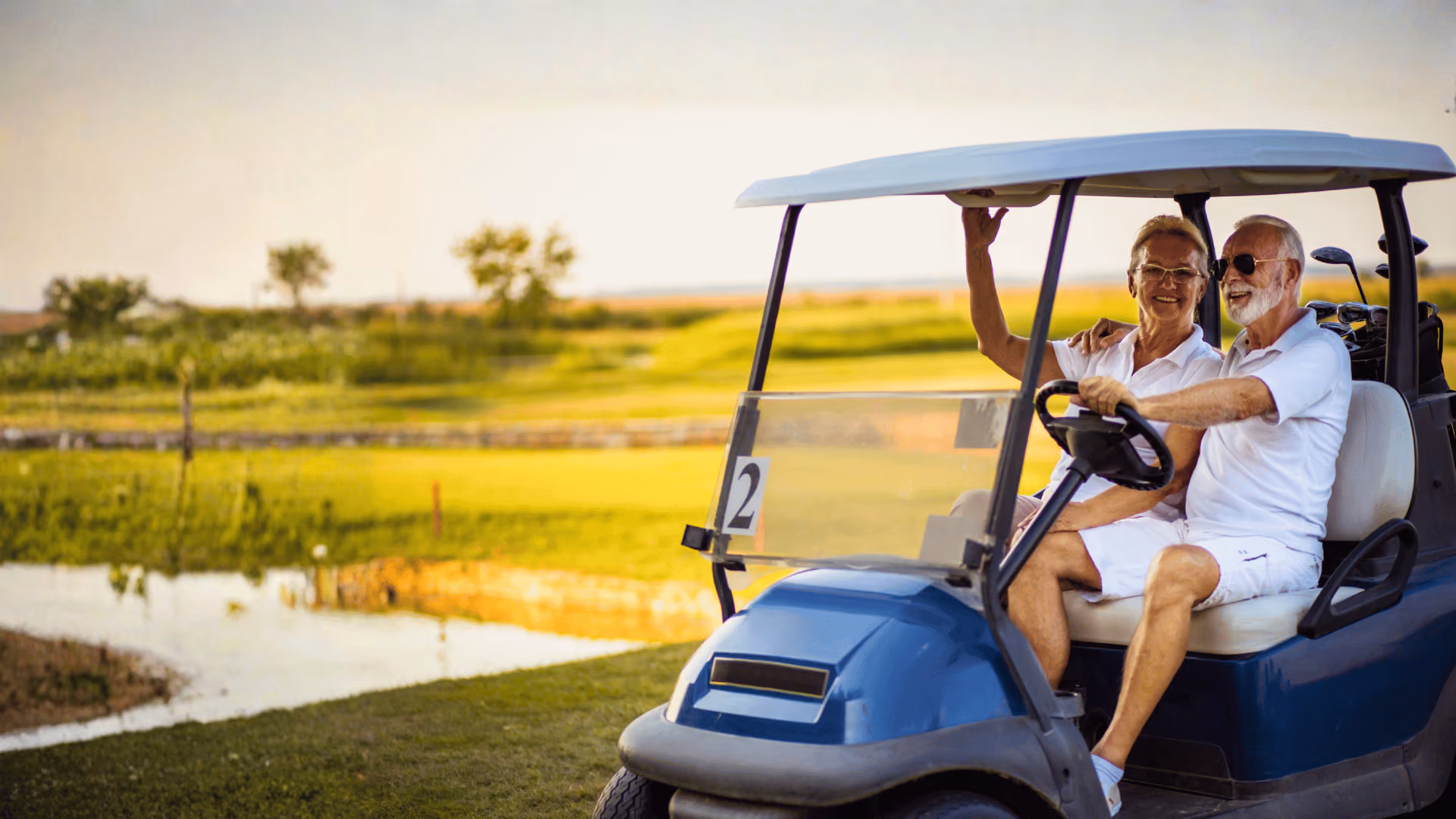 Smiling elderly couple sitting in a blue golf cart on a green golf course near a water hazard.