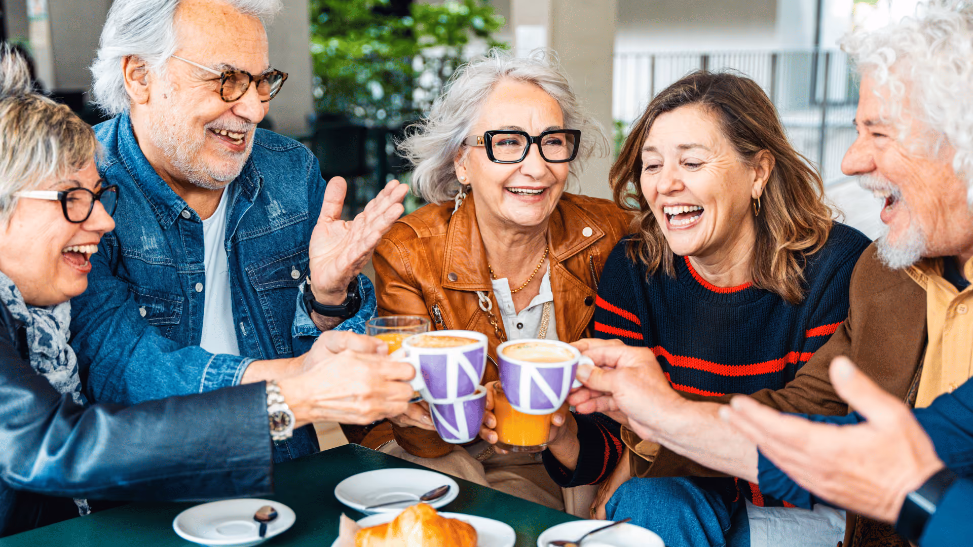 Five diverse older adults happily clinking coffee mugs and glasses of orange juice at a café table.
