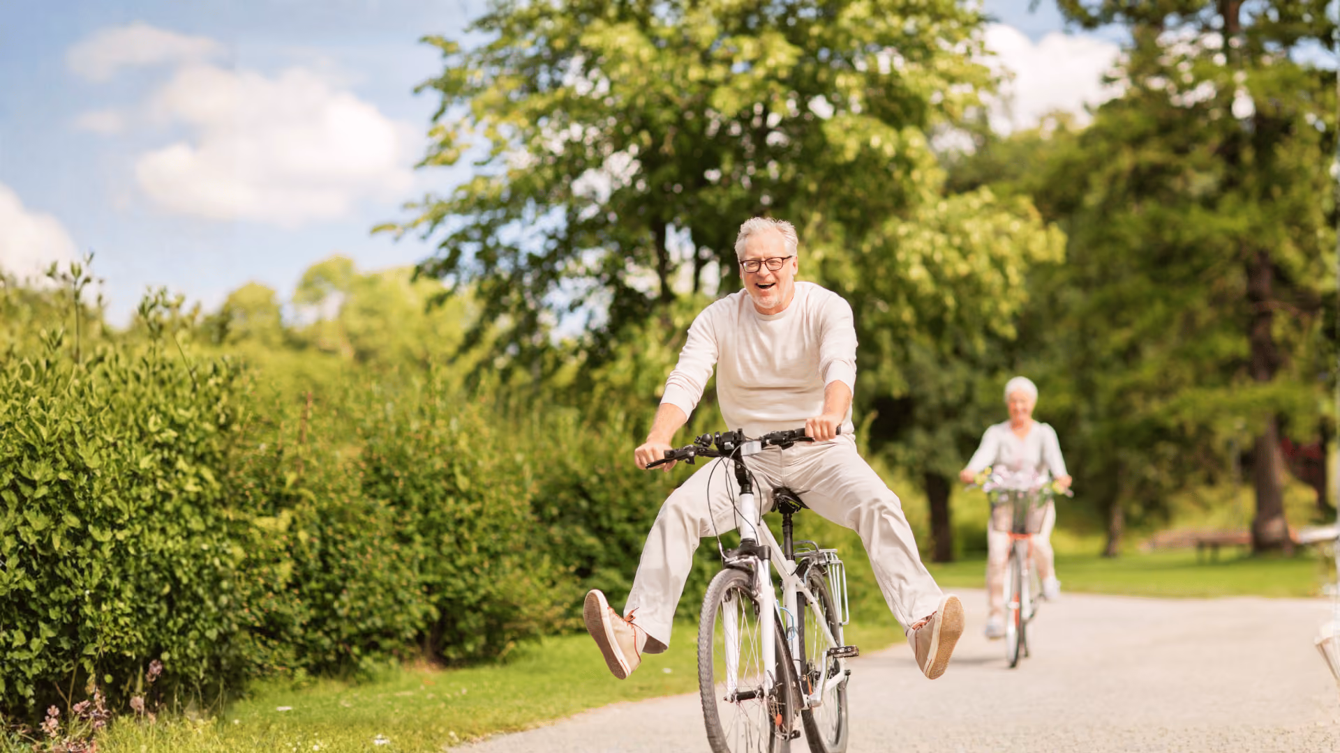 Elderly man joyfully riding a bike with legs out, woman cycling behind on a park path.