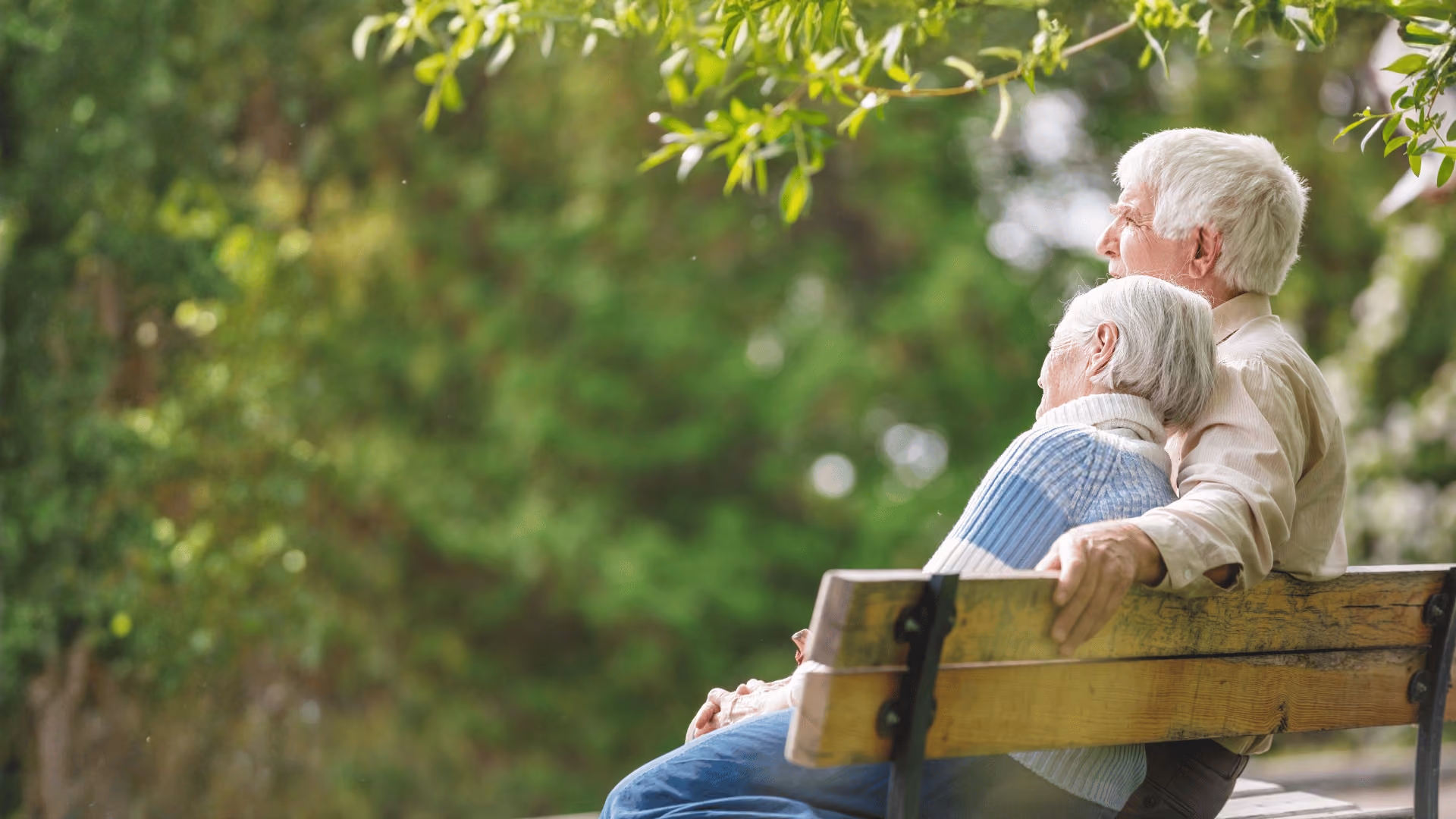 Elderly couple sitting on a park bench, leaning on each other, surrounded by green foliage.