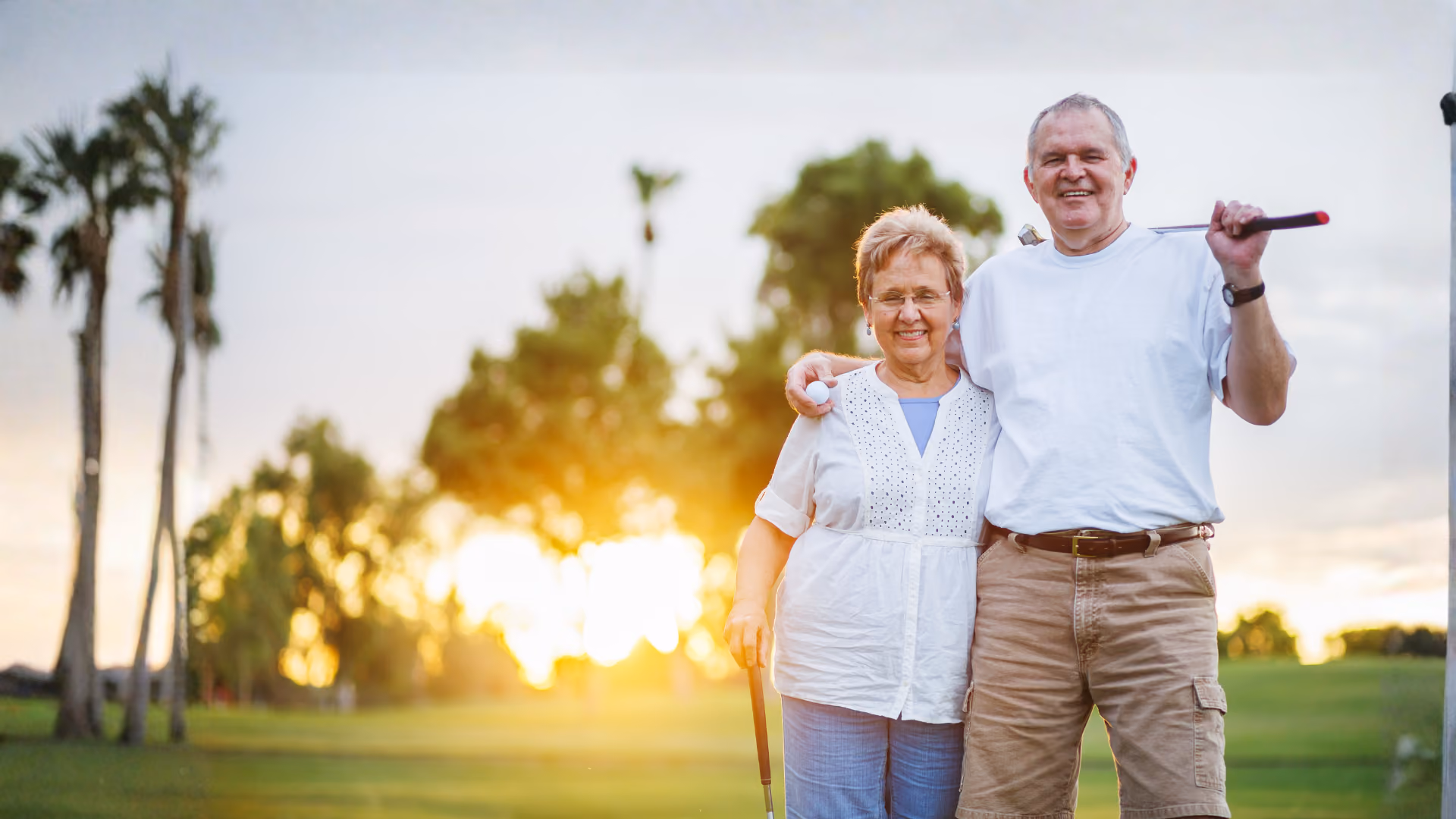 Smiling elderly couple standing on golf course at sunset, each holding a golf club.