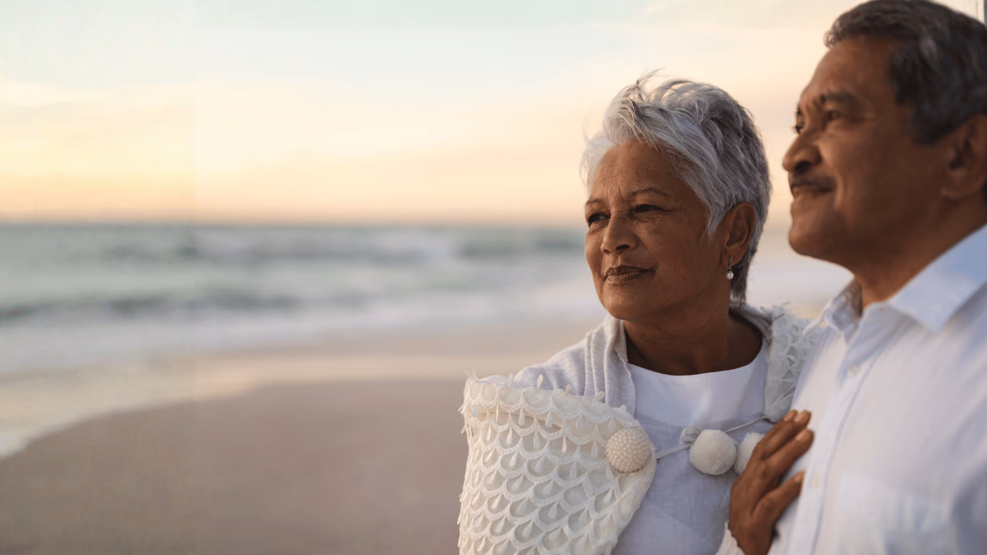 Older couple standing close on beach at sunset, looking thoughtfully into the distance.
