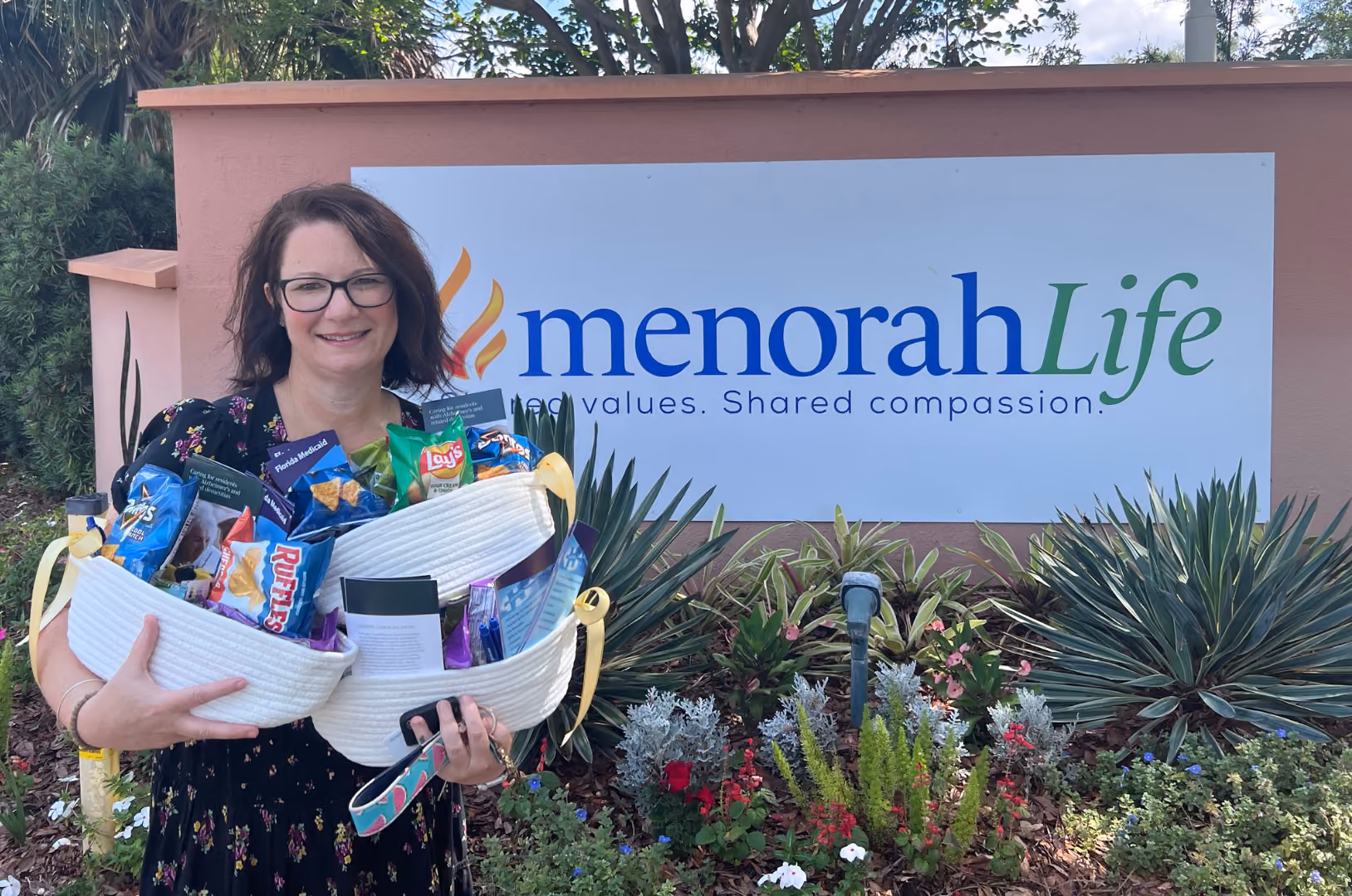 Woman holding baskets filled with snacks stands in front of a Menorah Life sign and garden.