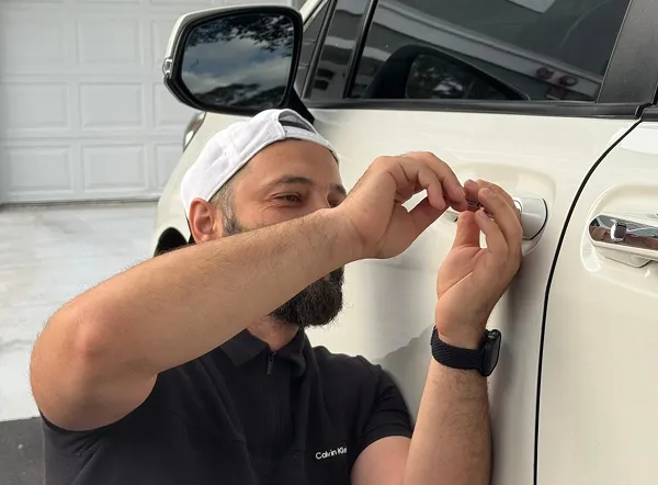 Locksmith technician unlocking a white car door with a tool on a driveway.