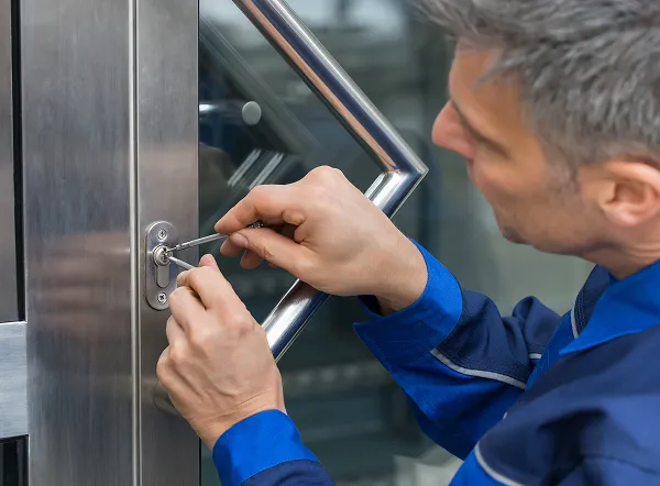A locksmith in blue uniform using tools to pick a lock on a metal and glass door.