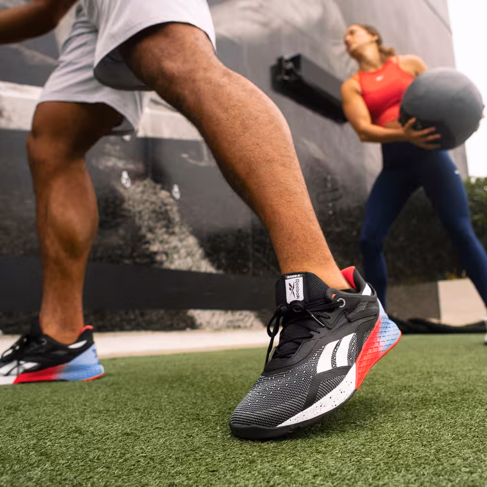 Close-up of the Reebok Nano X shoe in motion, with a female athlete holding a medicine ball in the background.