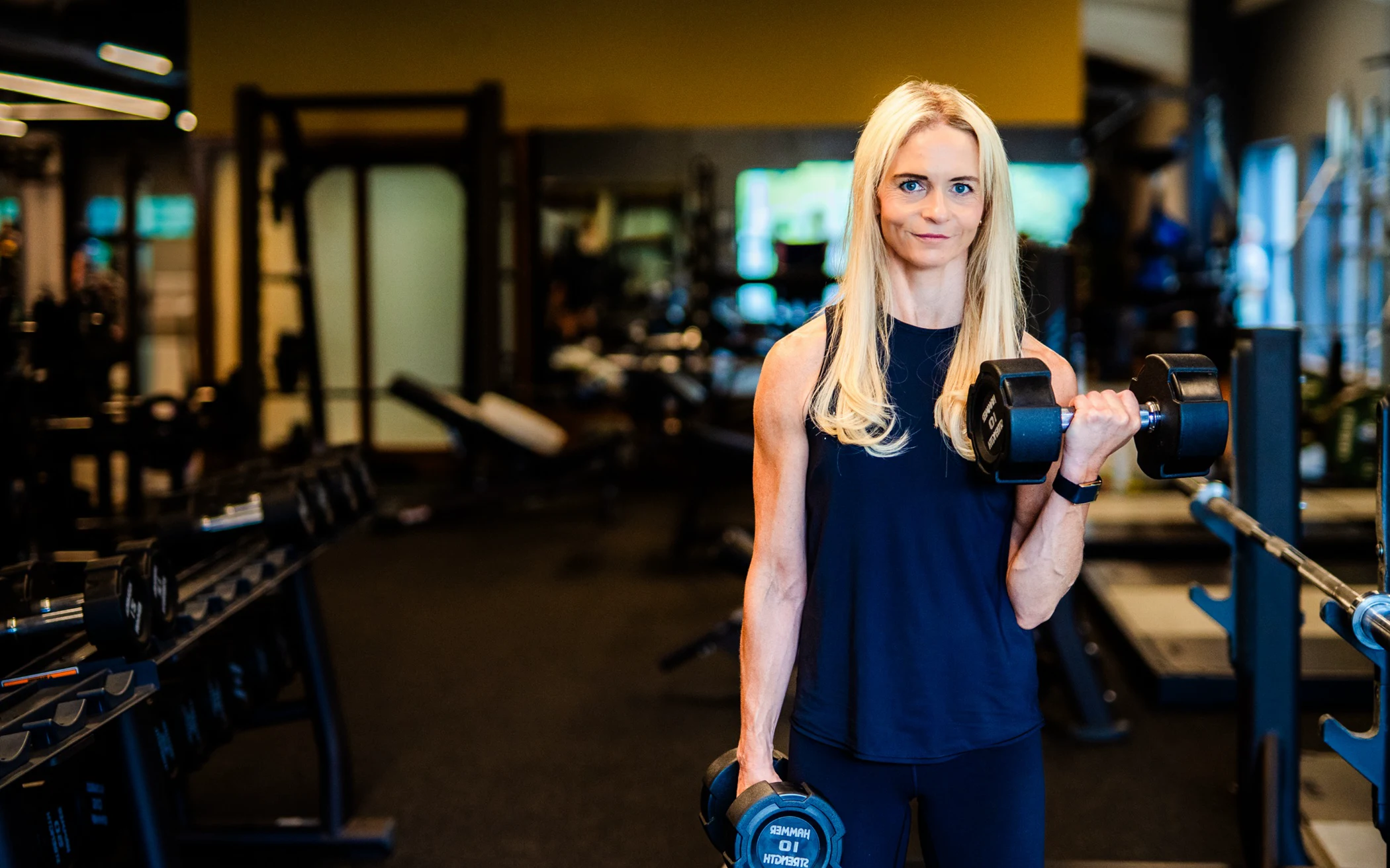 Woman with long blond hair lifting two dumbbells in a gym with fitness equipment around.