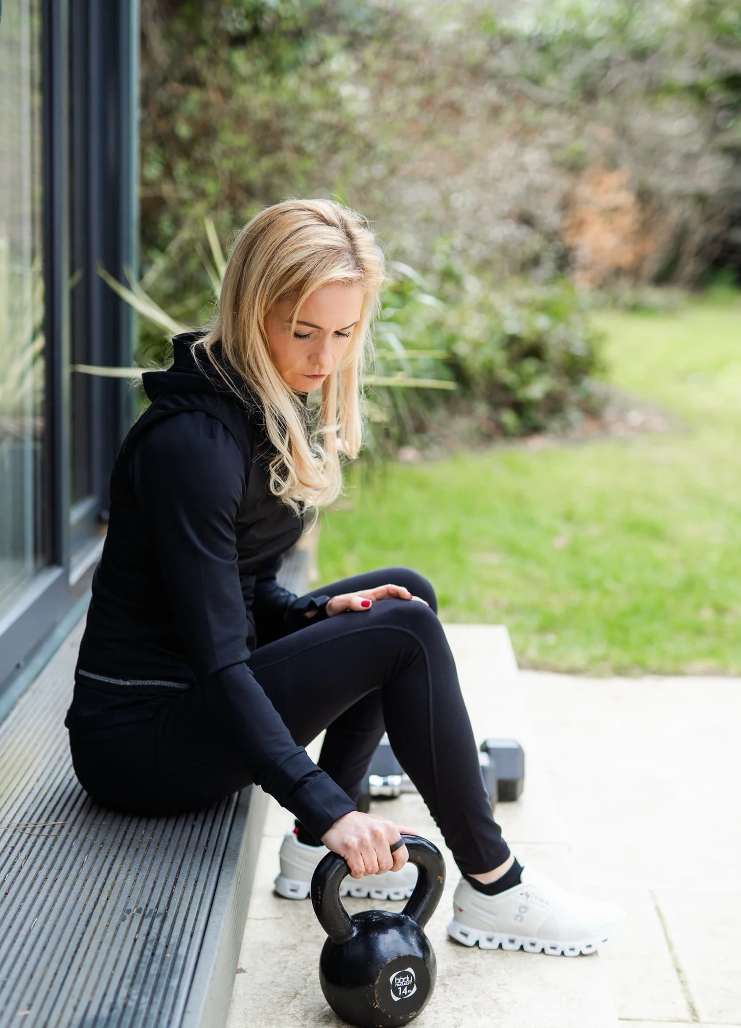 Woman in black workout clothes sitting outside holding a 14kg kettlebell near white shoes.