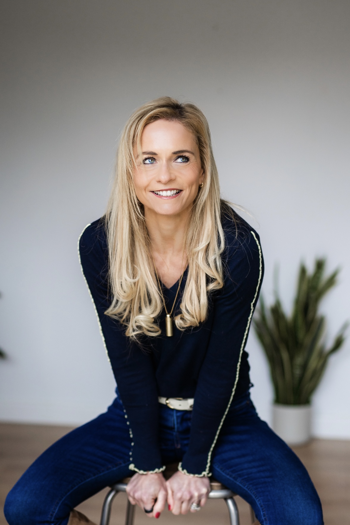 Smiling blond woman in black top and jeans sitting on a stool with indoor plants in background.