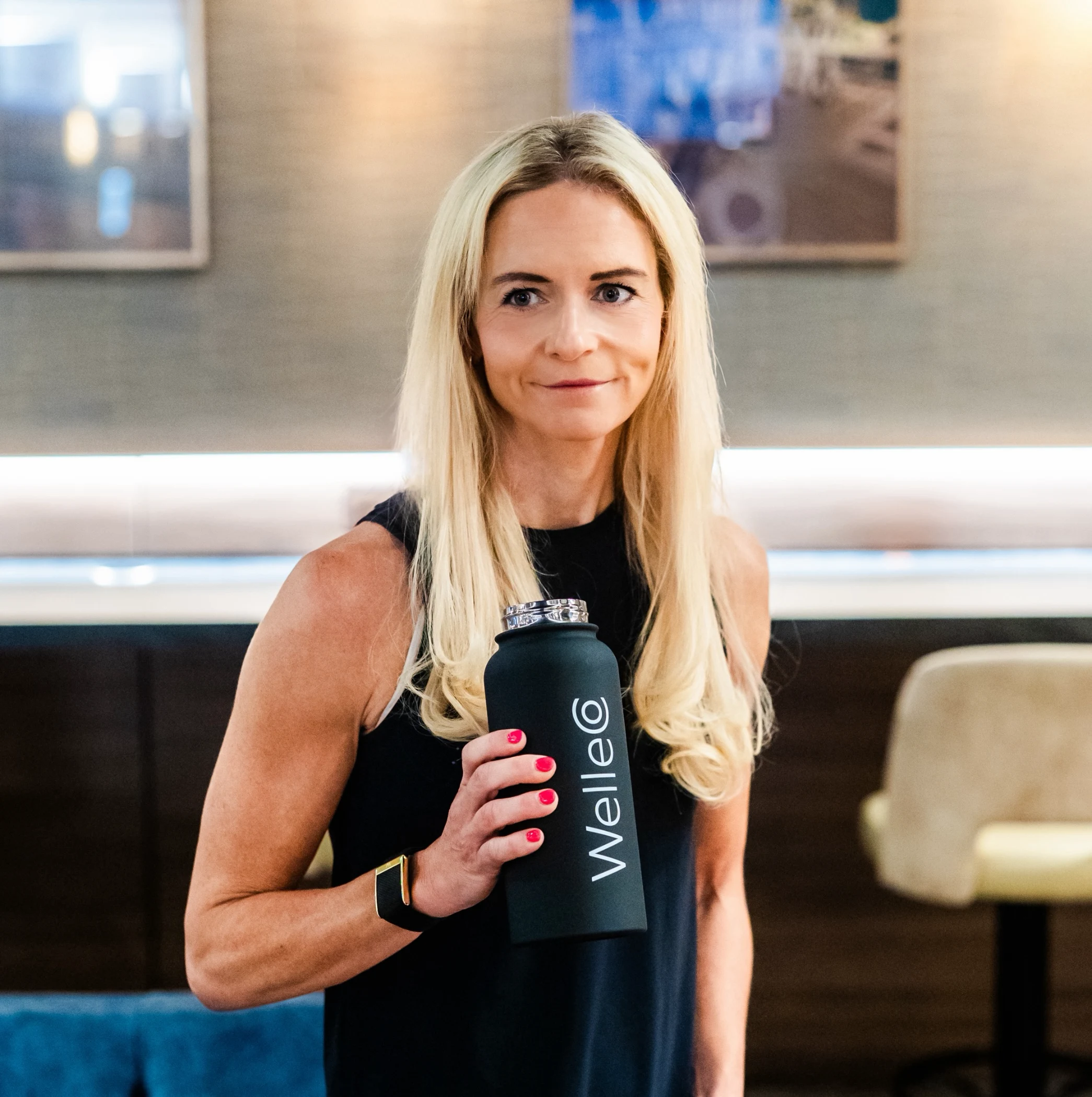 Blonde woman in black sleeveless top holding a black Welleco water bottle indoors.