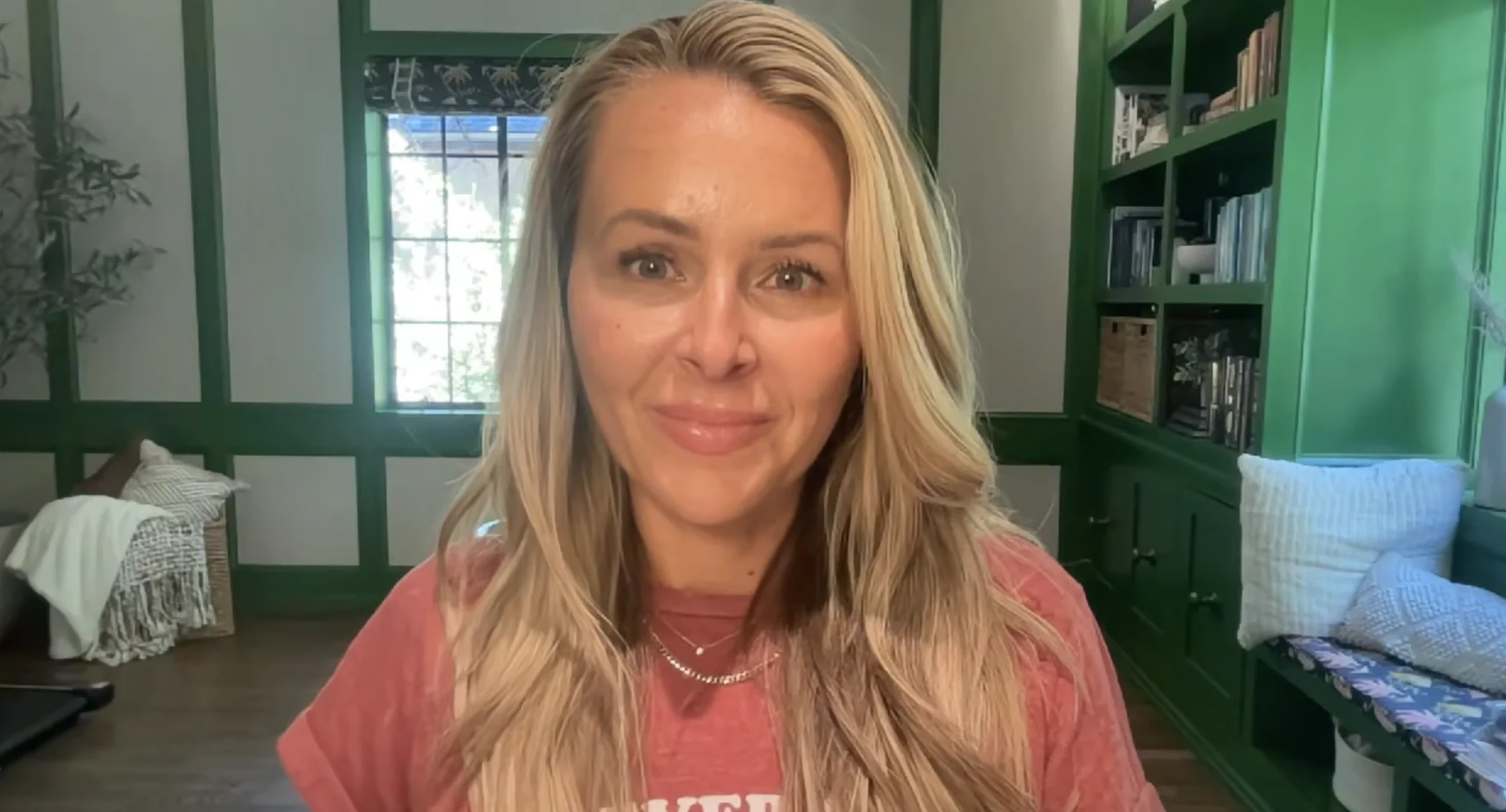 Smiling blonde woman with long hair wearing a red shirt, sitting in a cozy room with green shelves and window.