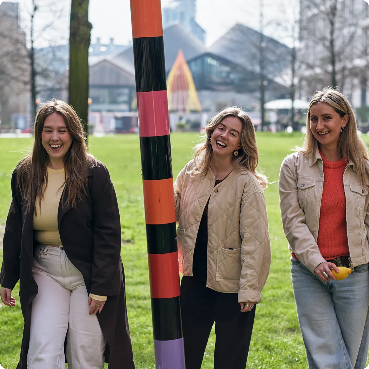 Three women smiling and standing outdoors with green grass and buildings in the background.