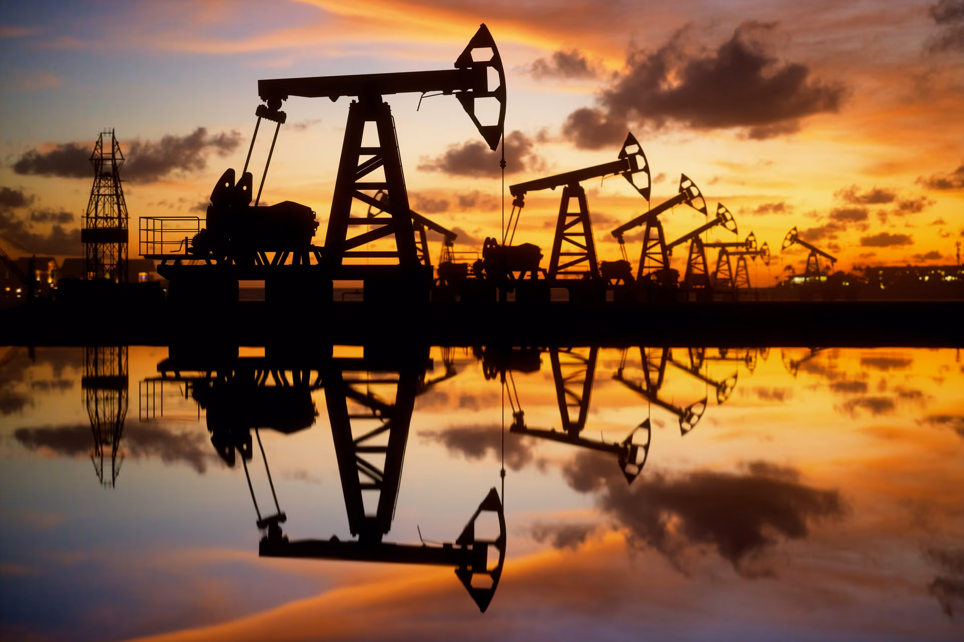 Silhouettes of multiple oil pump jacks reflected in water at sunset with colorful sky and clouds.