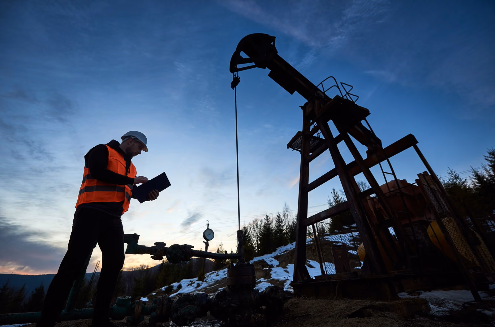 Worker in orange safety vest and hard hat checks clipboard by an oil pumpjack at dusk.
