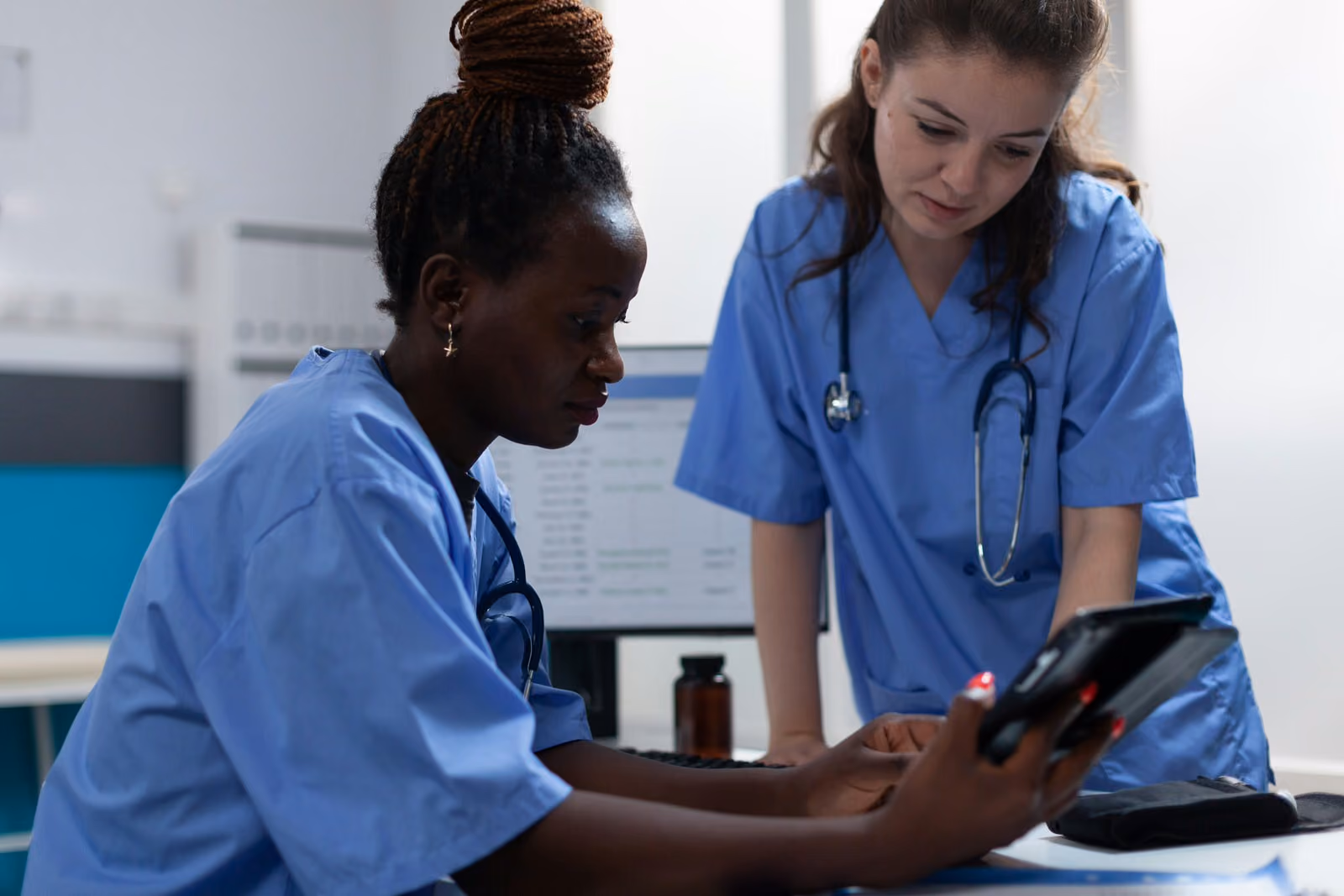 Two nurses in blue scrubs reviewing information on a tablet in a medical office.