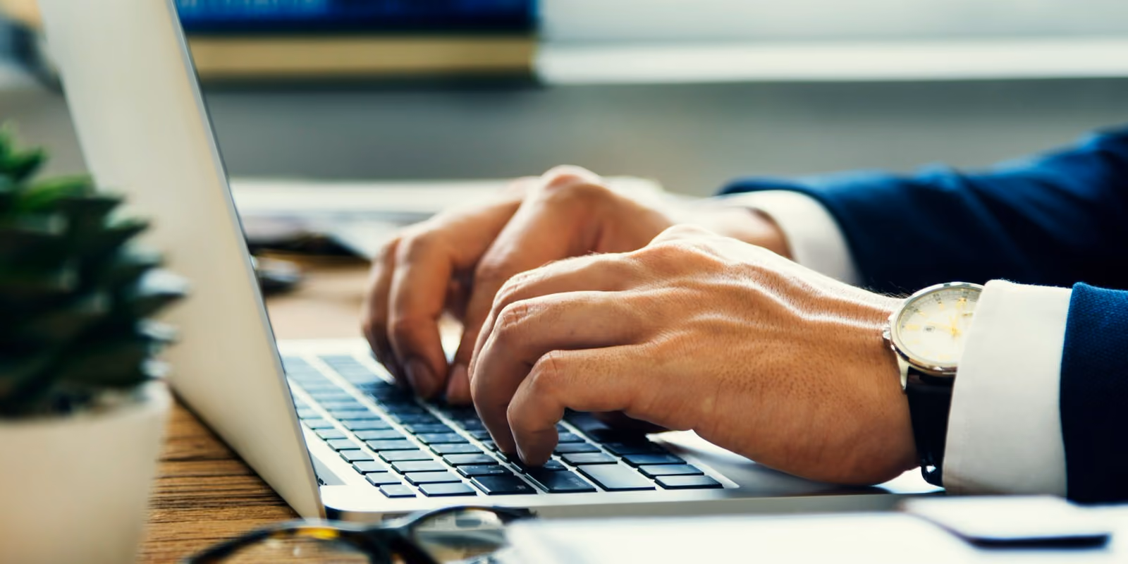 Close-up of hands typing on a laptop keyboard with a watch on the wrist in an office setting.