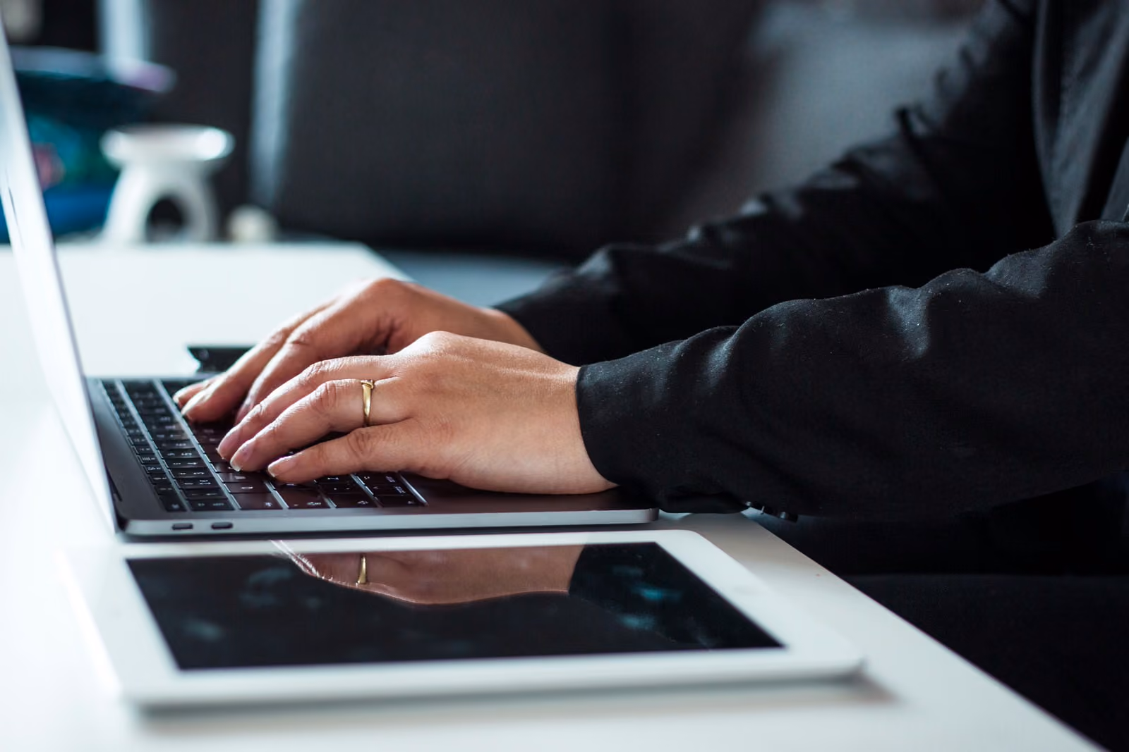 Person wearing a ring typing on a laptop keyboard beside a tablet on a white desk.