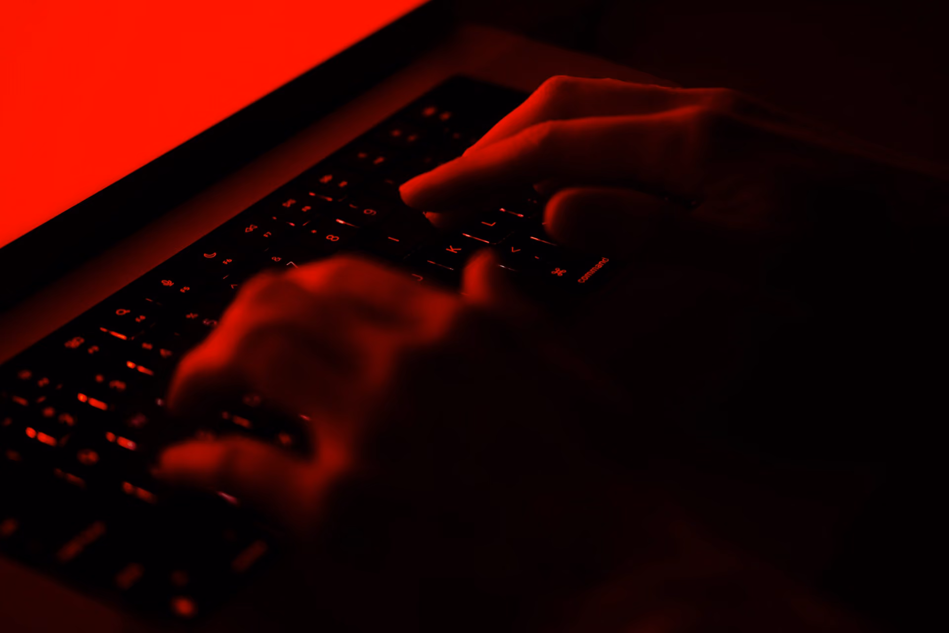 Hands typing on a keyboard with a red glow illuminating the keys and fingers in darkness.