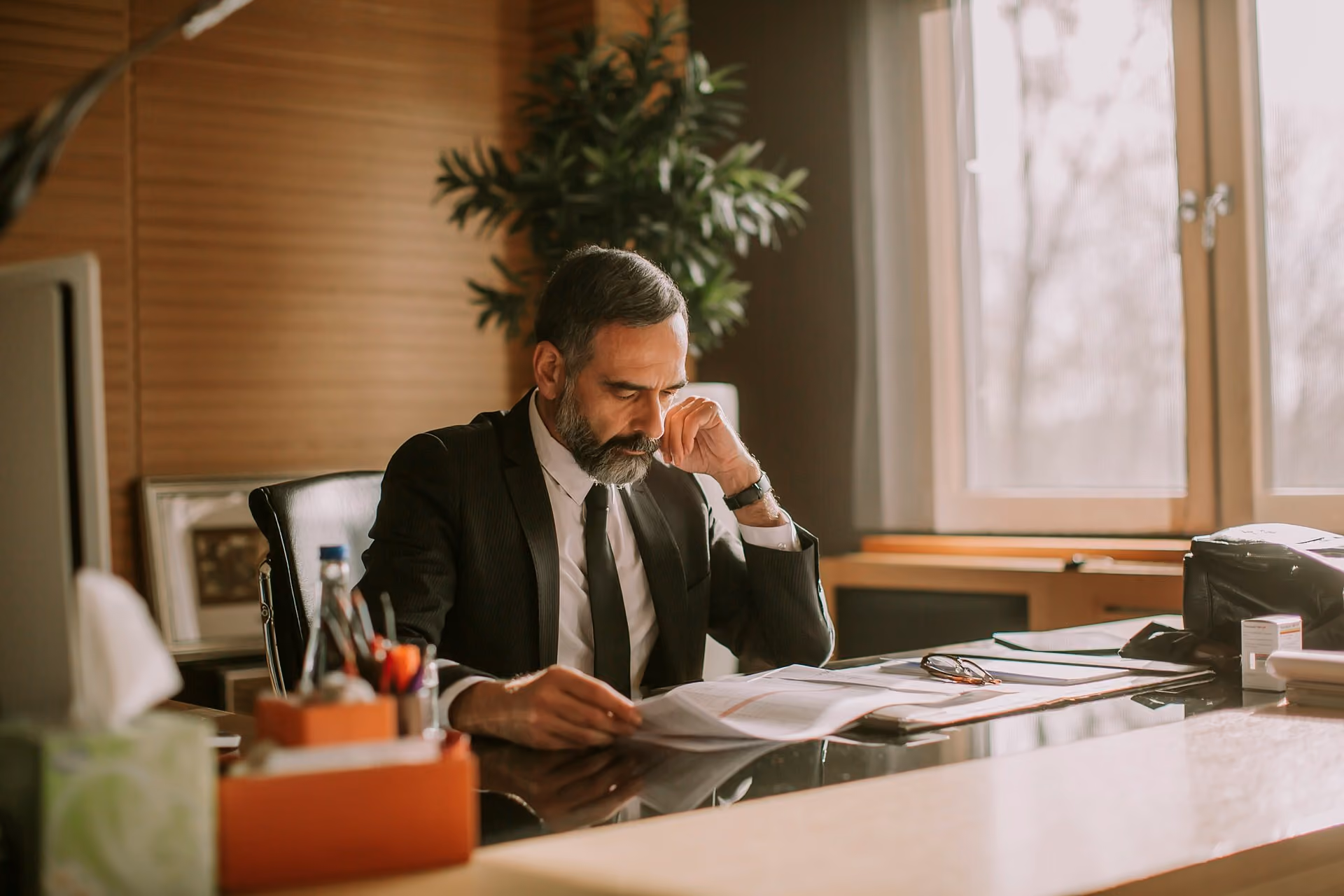 Mature man in suit reading documents at a desk in a warmly lit office with a large window.