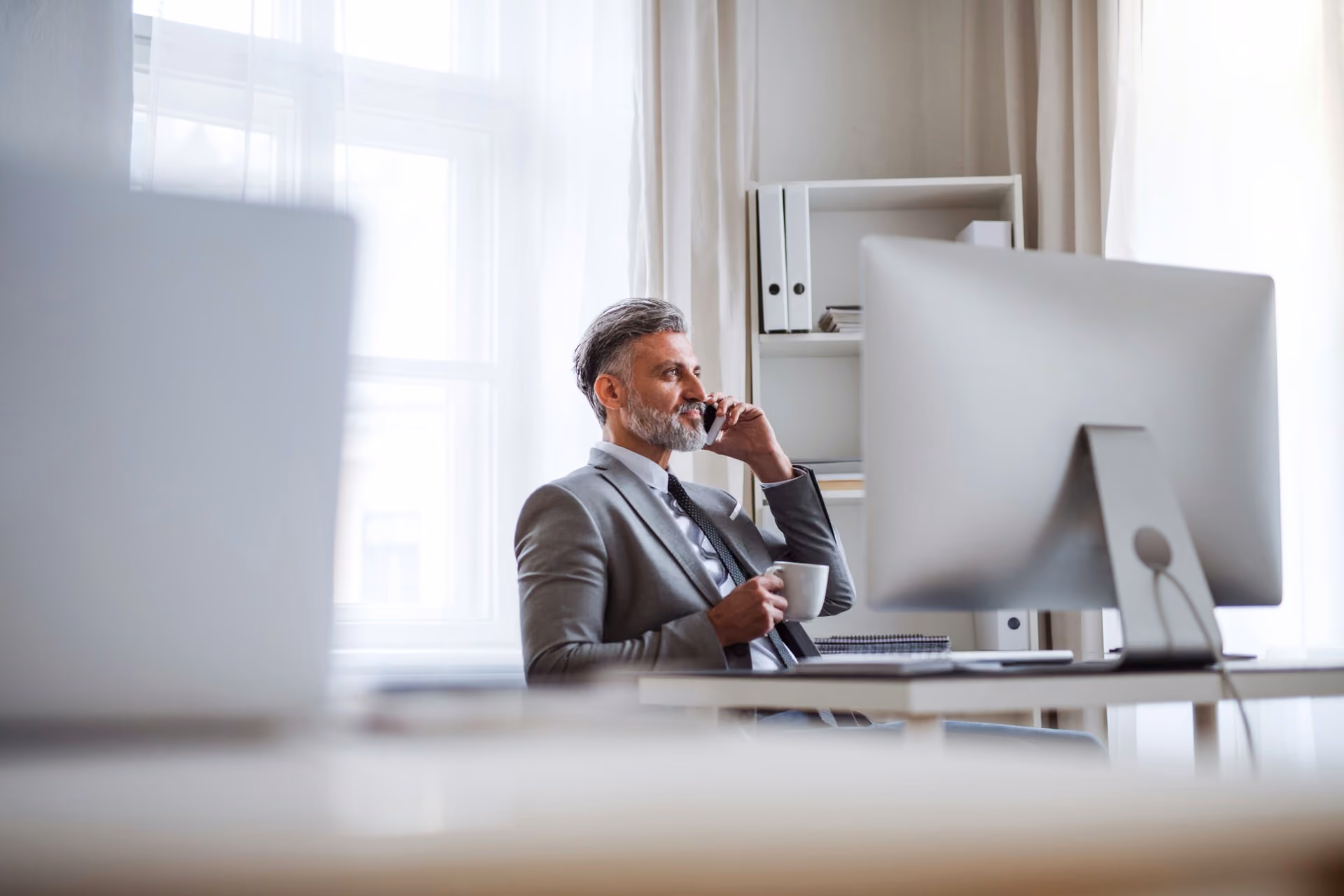 Middle-aged man in a suit sitting at a desk, holding a coffee cup and talking on a phone.