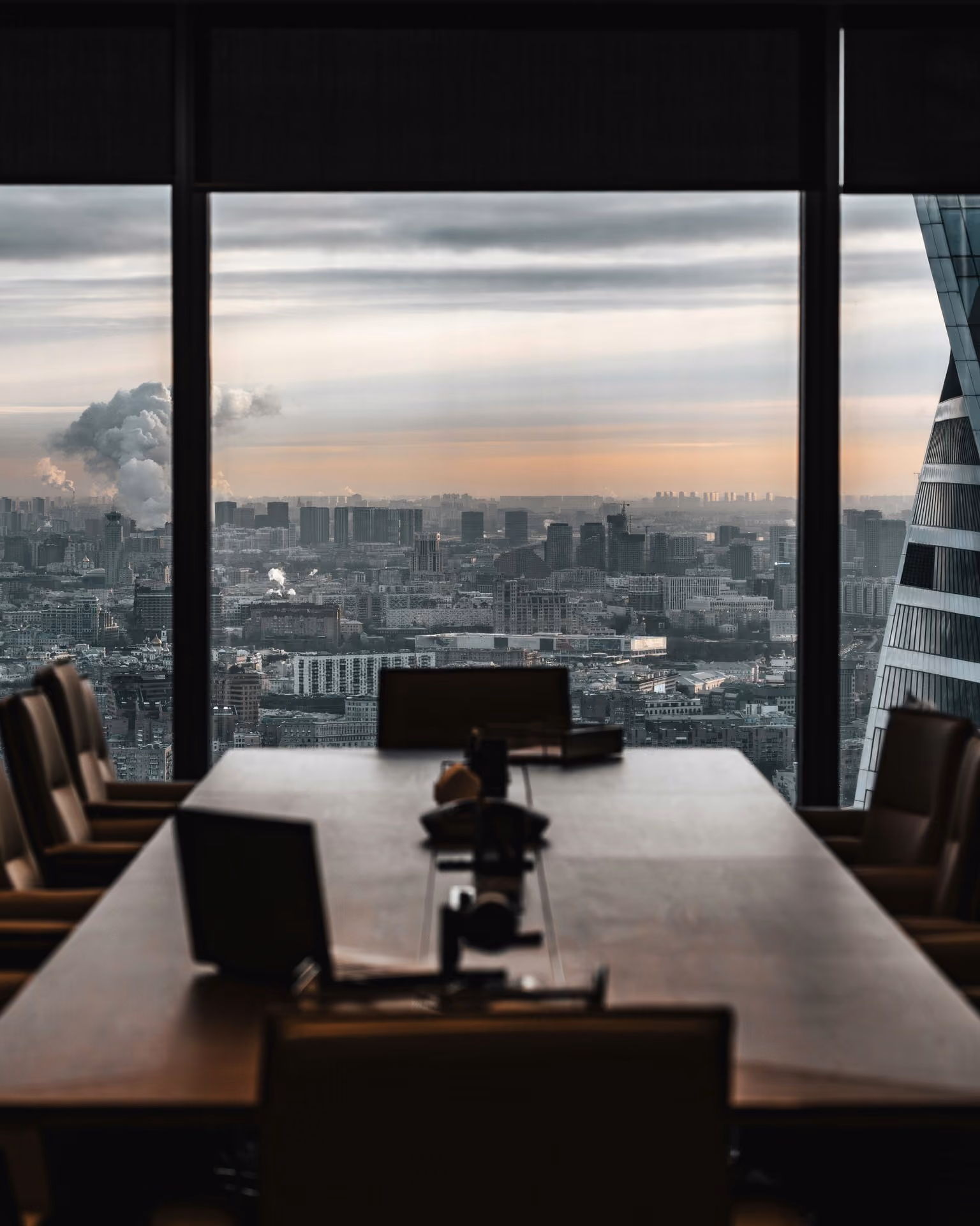 View of a cityscape with smoke from chimneys seen through large office windows behind a conference table.