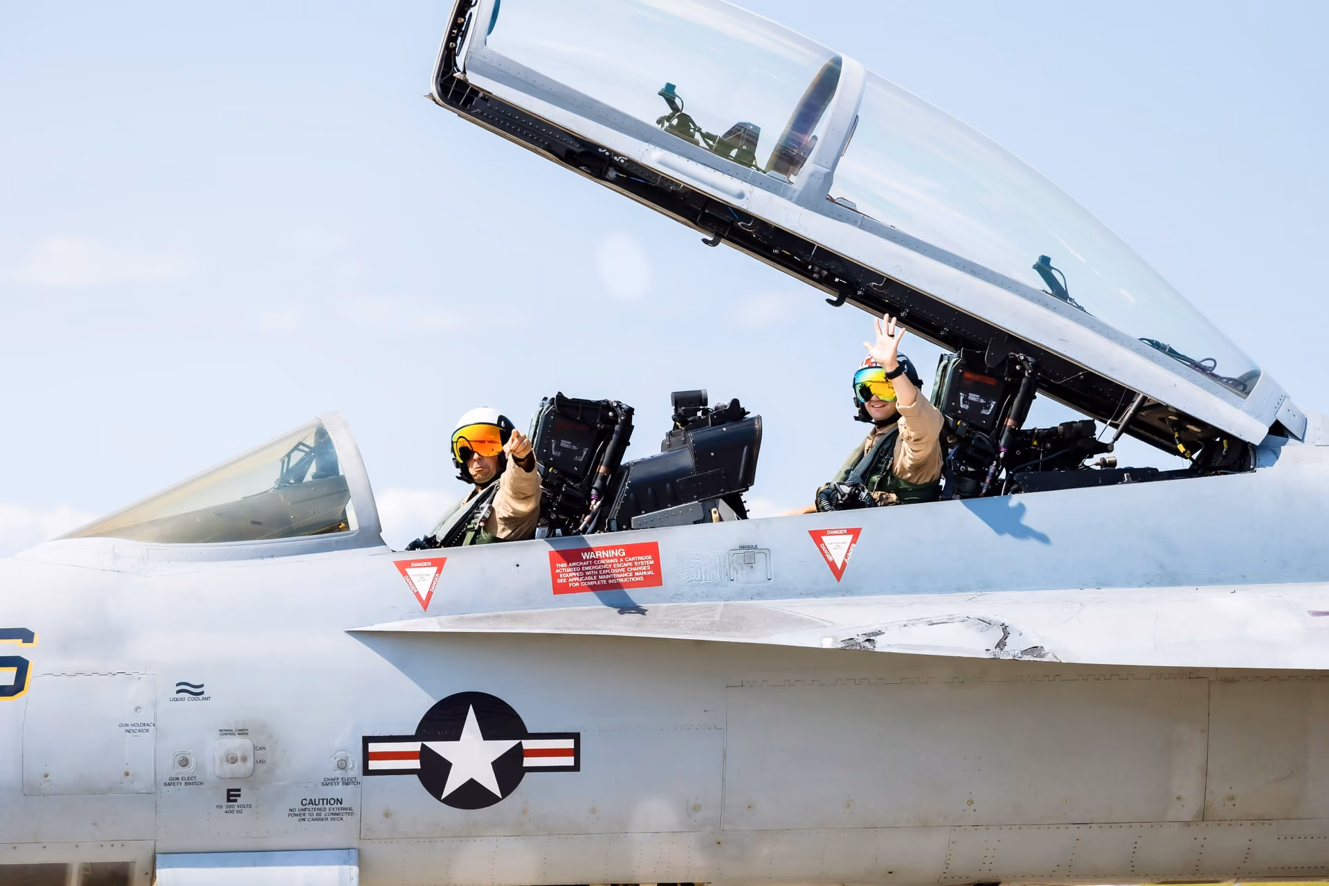Two pilots in helmets inside an open cockpit of a military jet, one waving and one pointing forward.