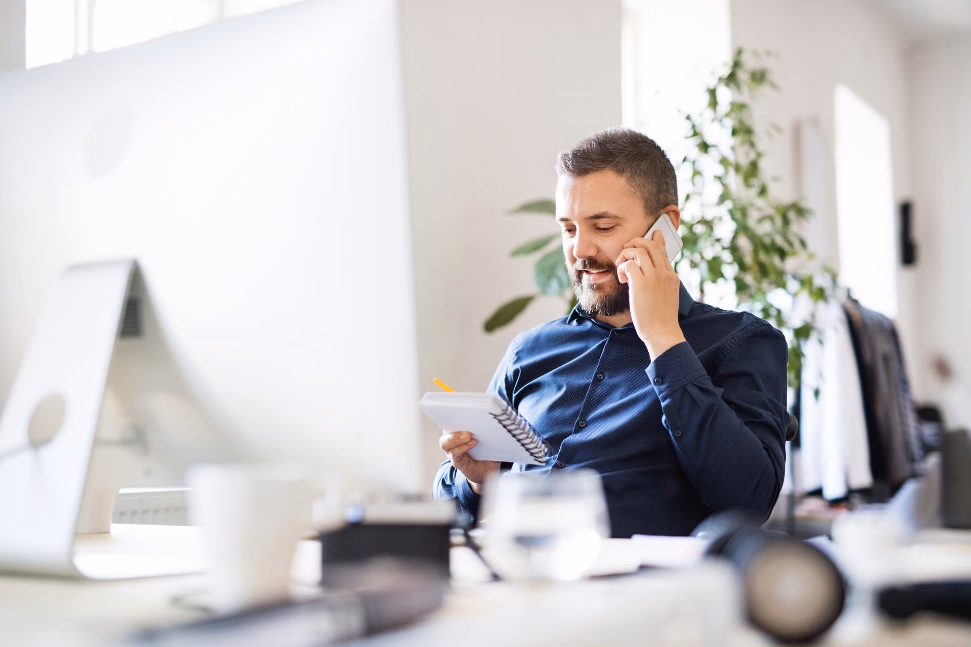 Man with beard in blue shirt talking on phone and writing in notebook at a bright office desk.