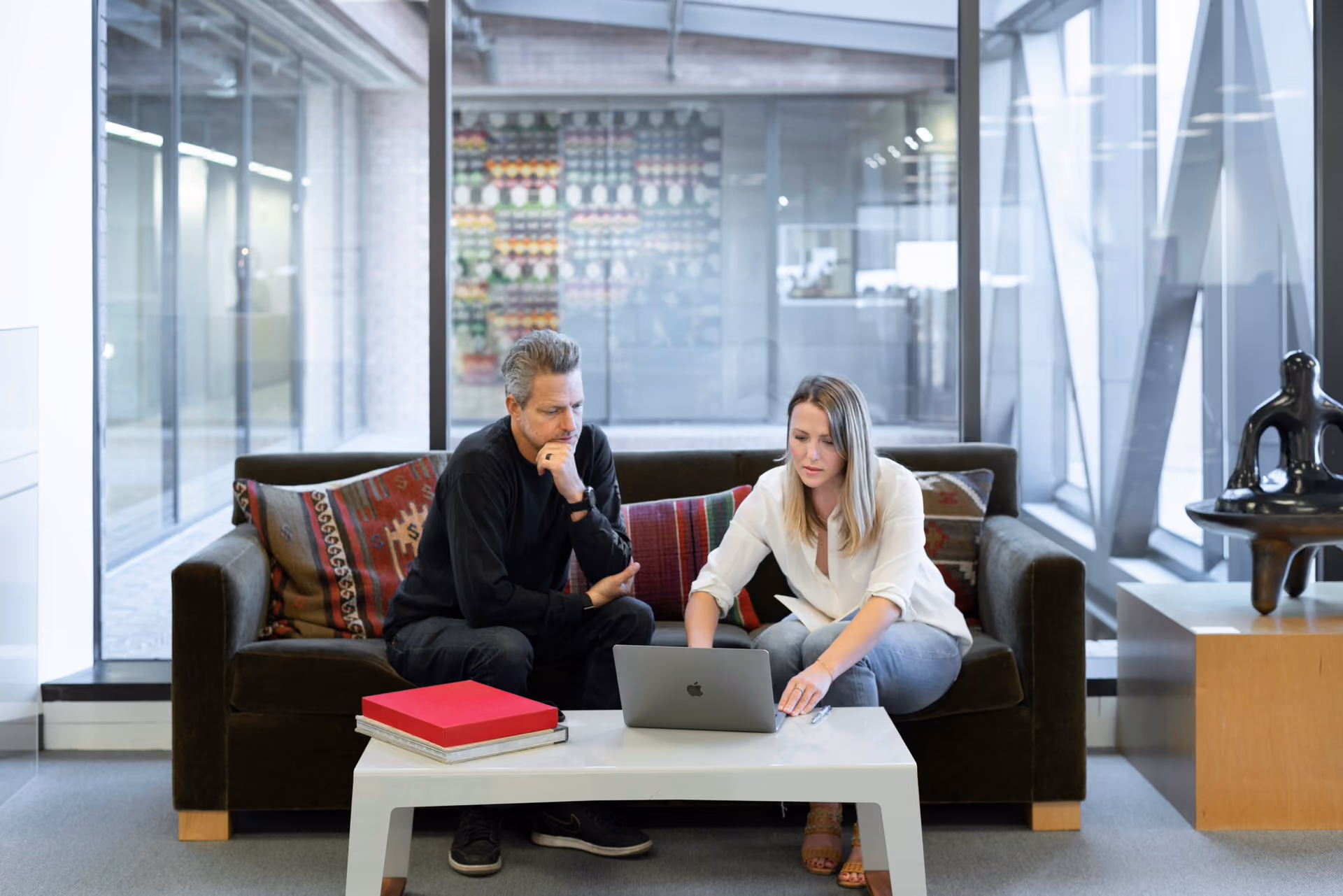 Man and woman sitting on a couch looking at a laptop on a white coffee table in a modern office.