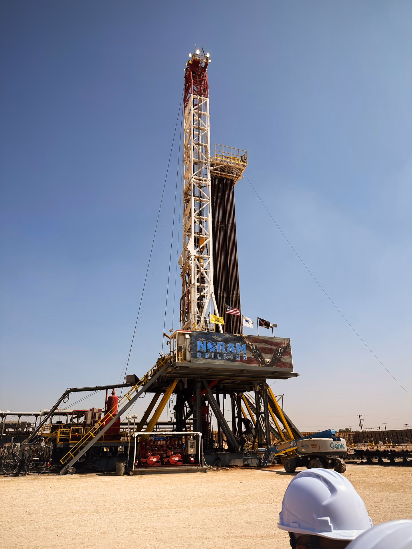 Tall Noram drilling rig with flags on a clear day, with workers wearing white helmets in the foreground.