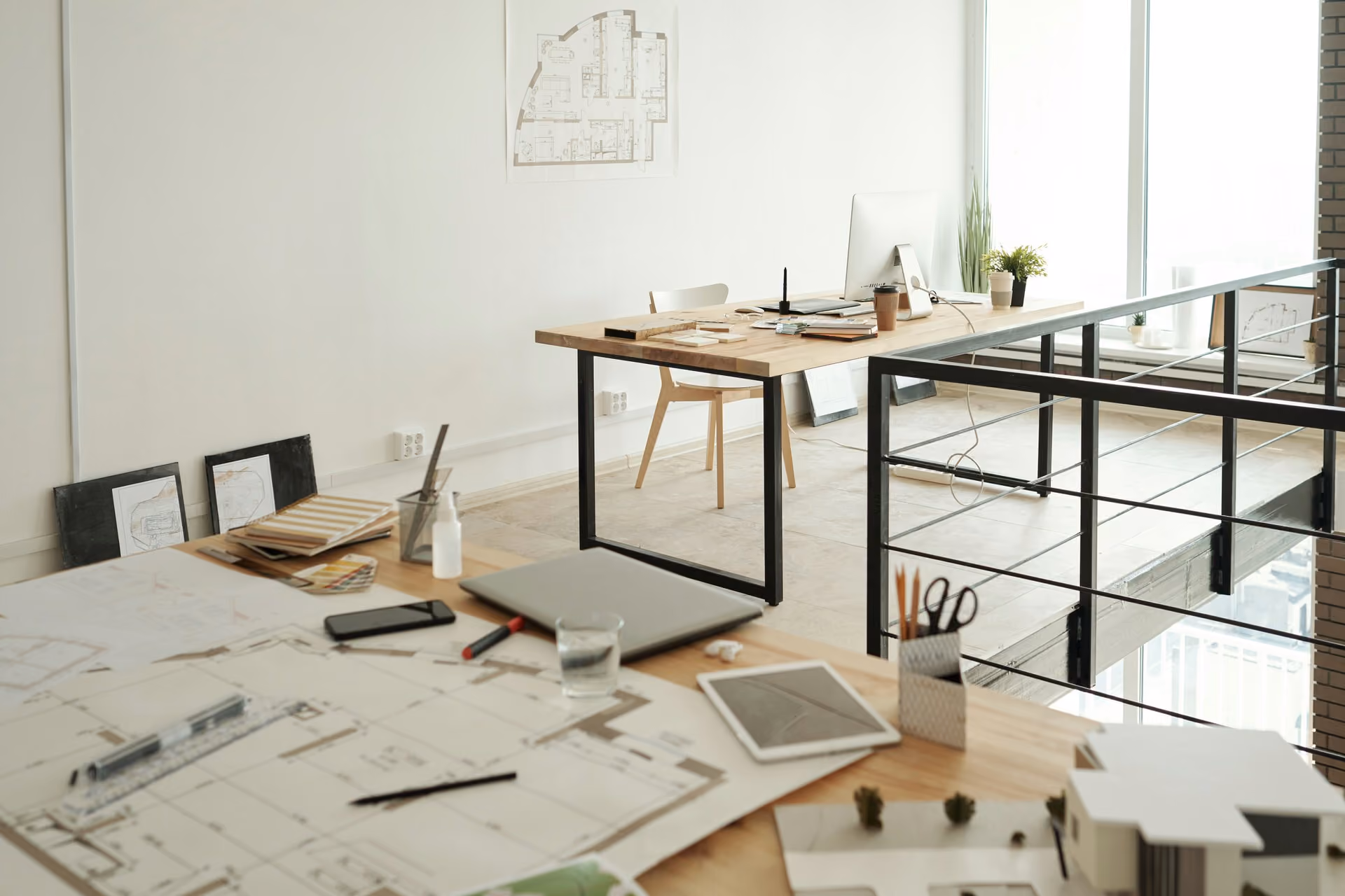 Architectural workspace with blueprints, model, and computer on wooden desks in a bright modern office.