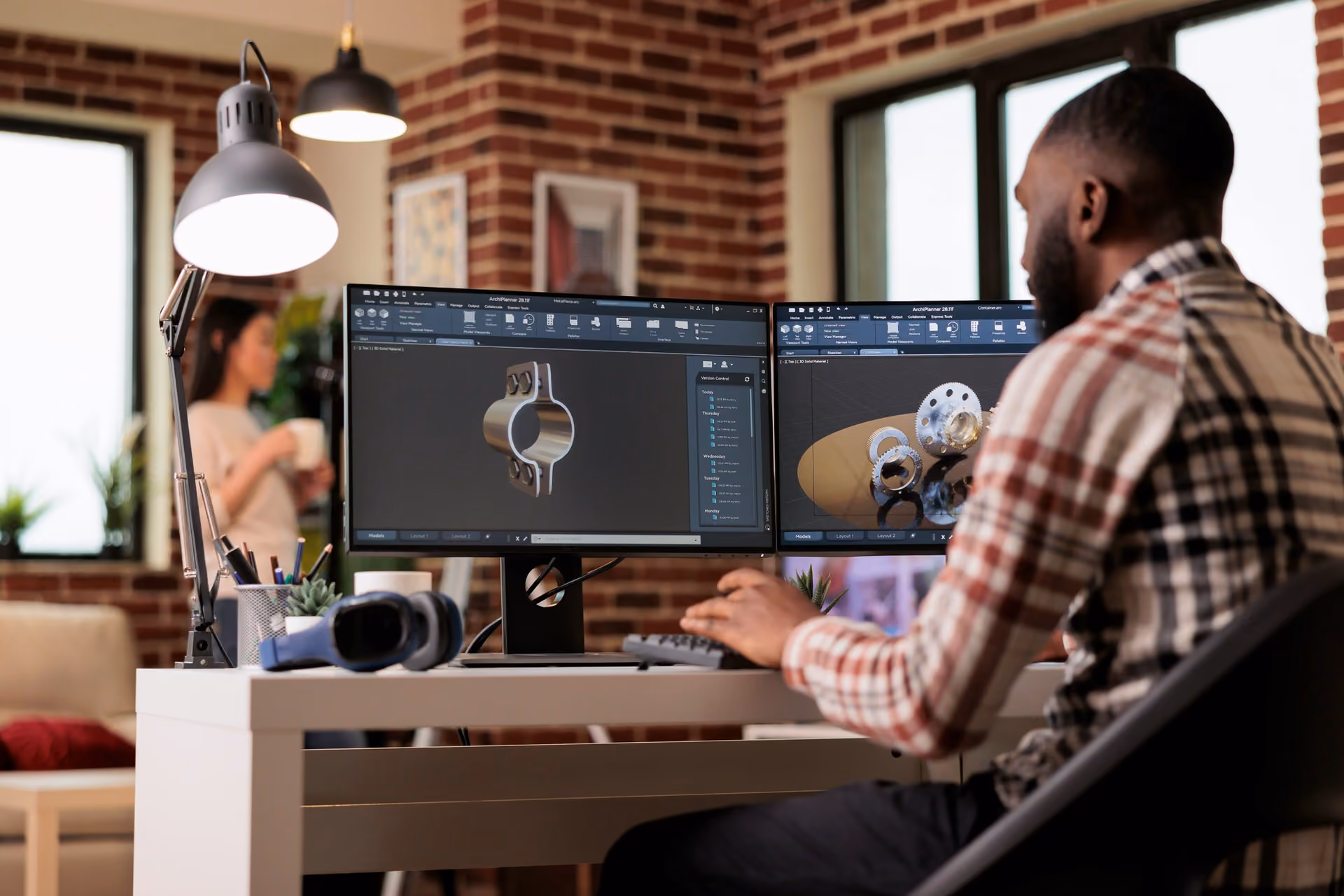 Man working on dual monitors with 3D mechanical part designs in an office with brick walls.