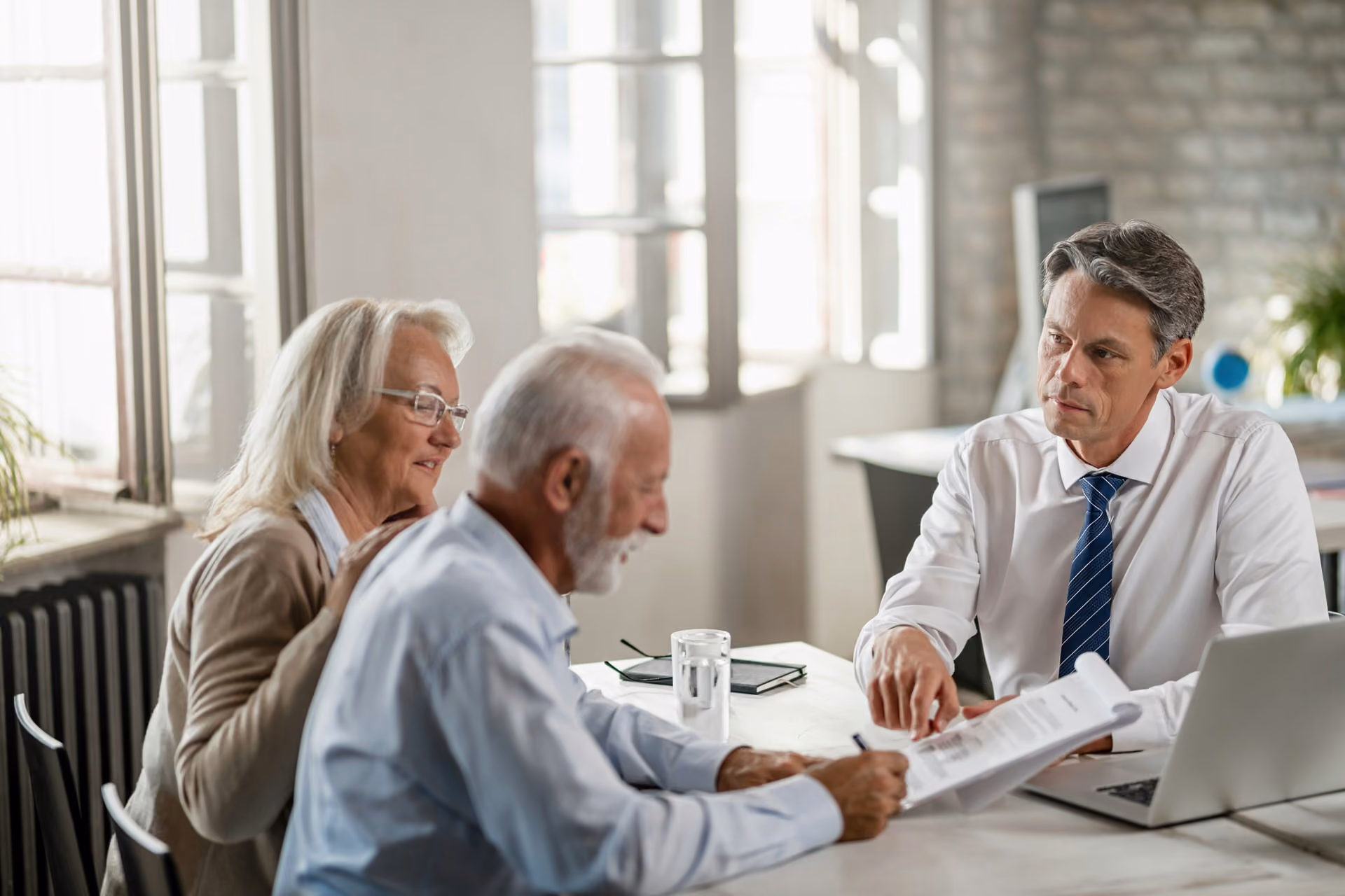 Older couple signing documents while man in shirt and tie reviews papers at office desk.