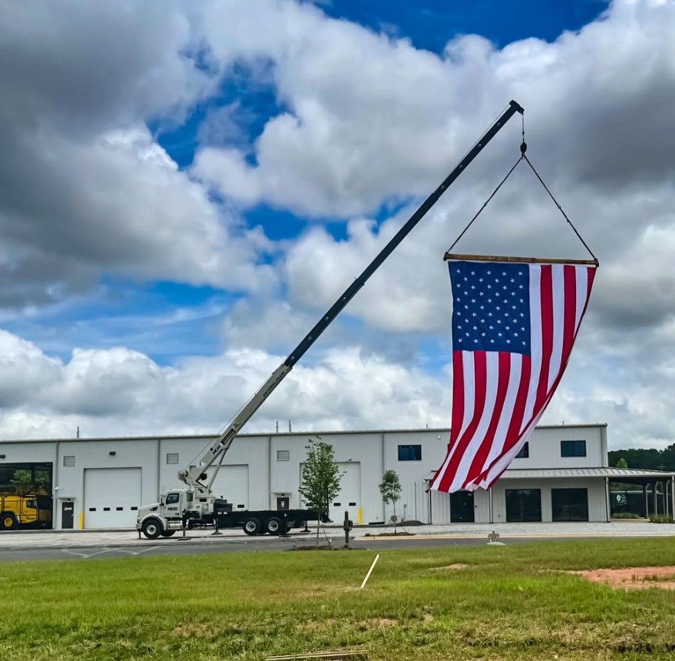Patriot Lifting truck-mounted boom crane flying a large American flag in front of an industrial warehouse