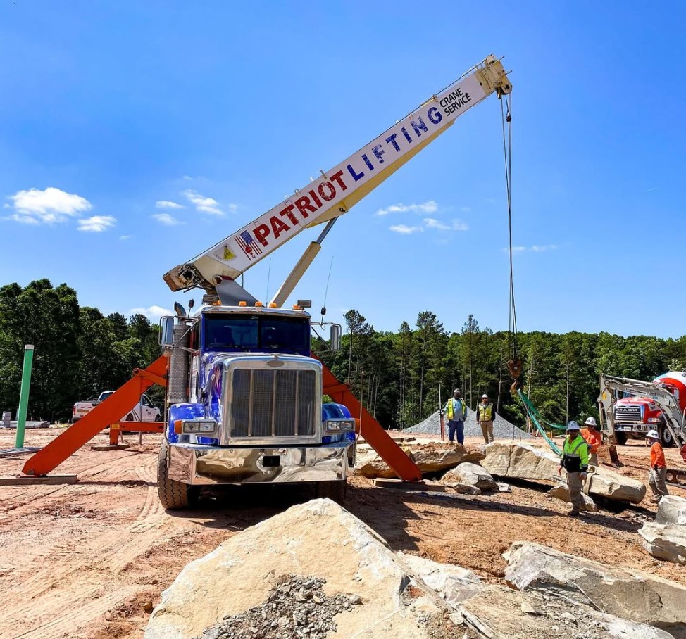 Patriot Lifting blue Peterbilt boom truck placing large landscape boulders with workers below
